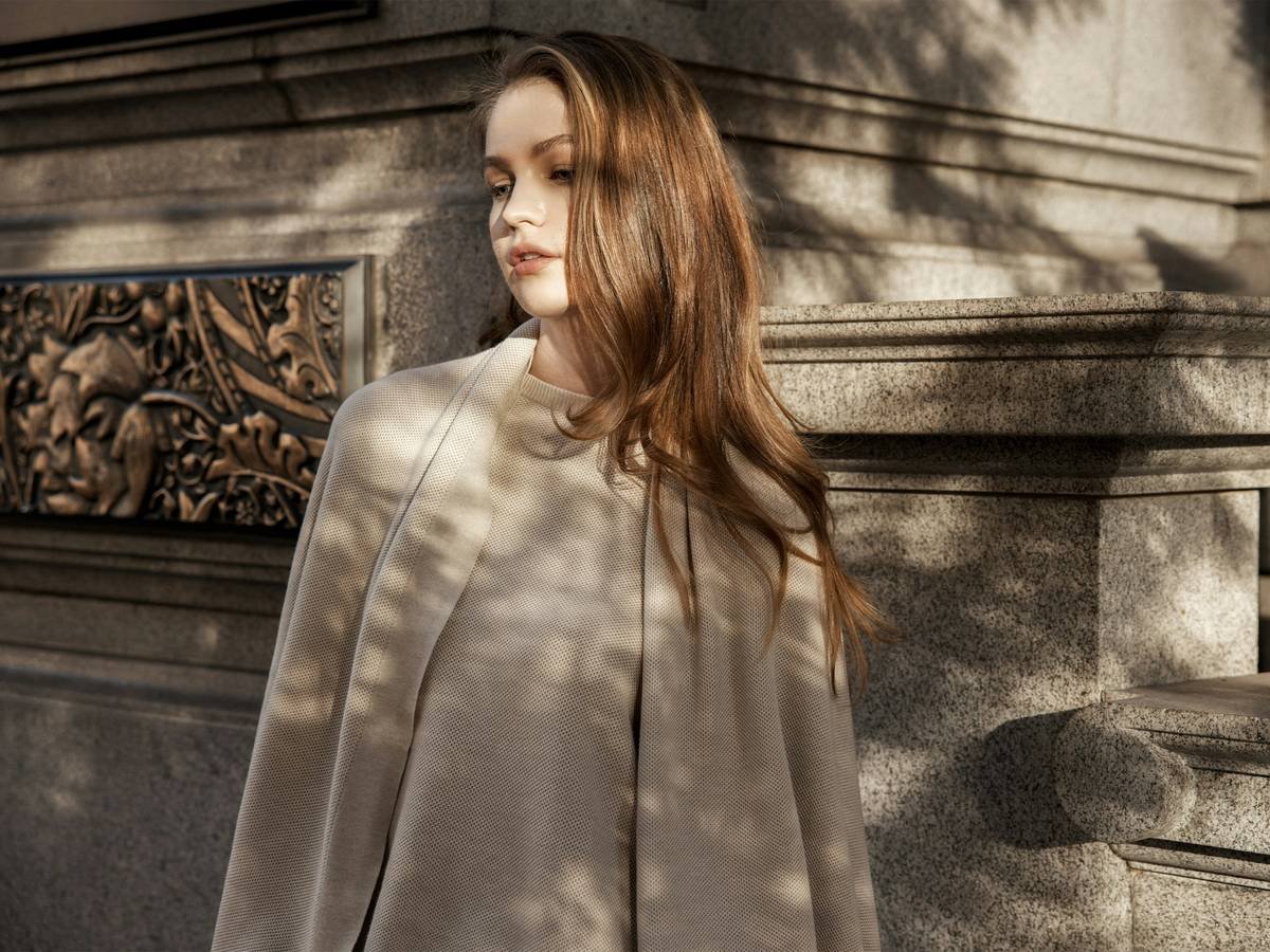 A woman stands by ornate stone architecture, wearing a neutral-toned wrap by MANITO for an understated, elegant look.