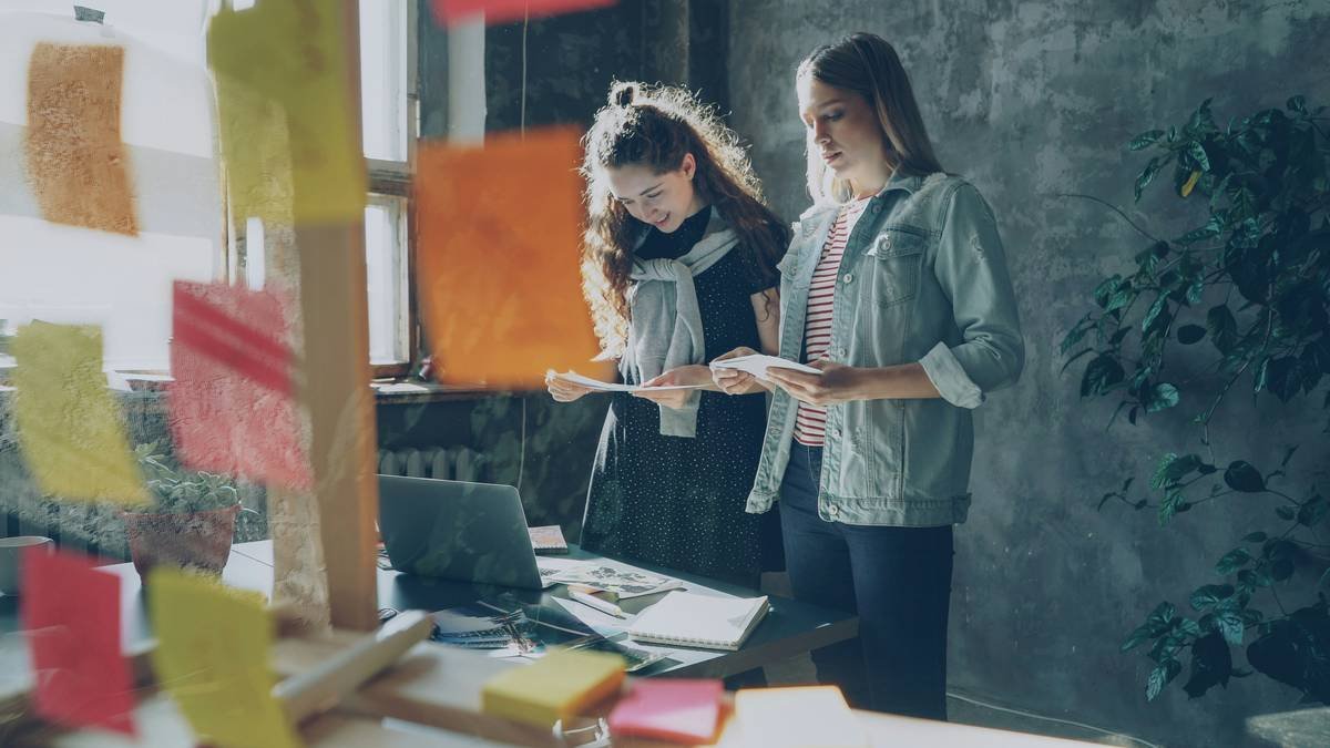 Cheerful designers are standing near table in loft style office and choosing photos for project. Young women are having friendly relations, they are chatting, laughing and sharing ideas.