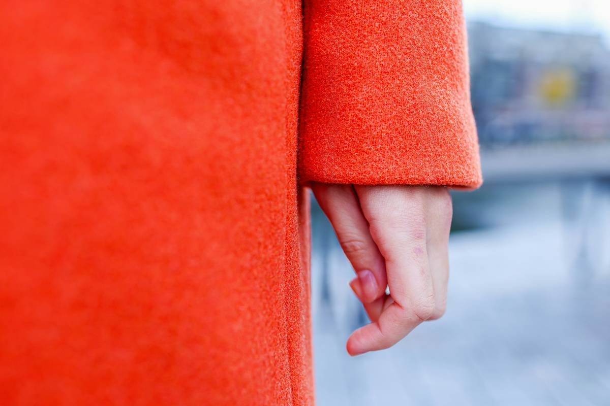 Minimalist fashion detail showing a relaxed hand emerging from the sleeve of a vibrant red-orange wool coat against a muted urban backdrop.