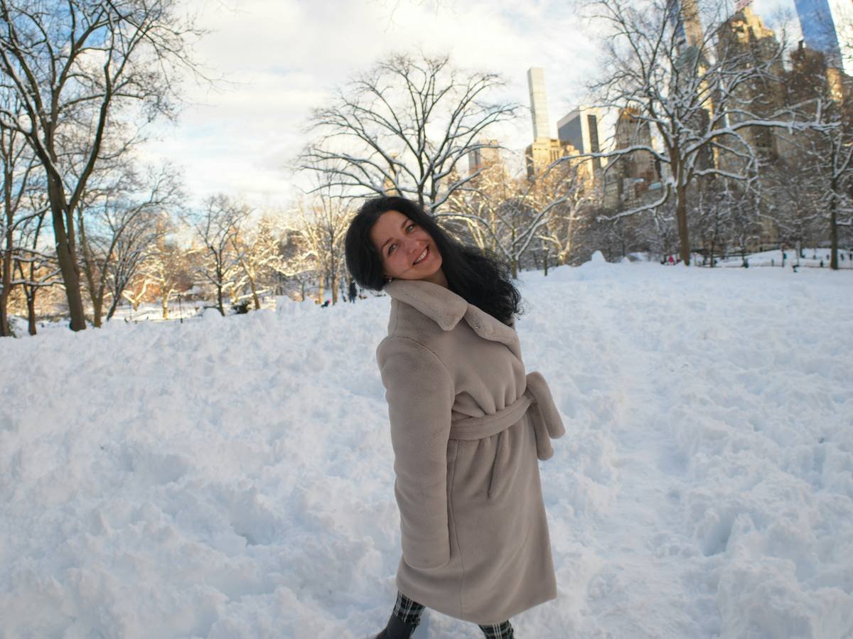 Photo by Olek Buzunov @buzunov.photoart. She is a Ukrainian singer living in NYC, captured during a personal winter photoshoot in Central Park. Surrounded by fresh snow and soft natural light, she interacts playfully with a snowman and poses among bare trees and city skyline views. The neutral tones of her coat contrast beautifully with the crisp white landscape, creating a calm, cozy atmosphere that blends urban life with seasonal serenity.
