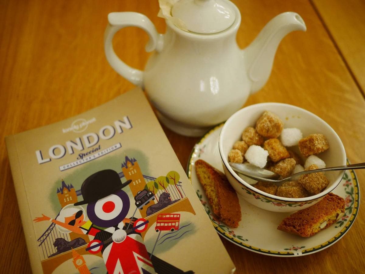 Photograph of a tea setting with a London travel guidebook, taken on location by the photographer. The image shows a white teapot, a bowl of sugar cubes, and biscuits arranged on a wooden table alongside an illustrated guide to London, evoking everyday British culture and quiet ritual rather than landmarks. The warm light and simple composition create a calm, domestic still life with cultural context.